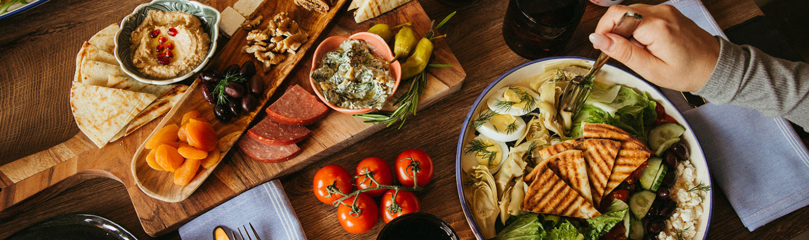 mezze items, tomatoes, and a cobb salad on a wooden table with person eating